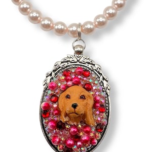 May include: A pendant necklace featuring a brown dog portrait within an oval frame, surrounded by pink and red crystals. The pendant hangs from a strand of light pink faux pearls.