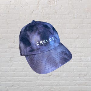 May include: A black and blue galaxy print baseball cap with the word "CRESCO" embroidered in white on the front.