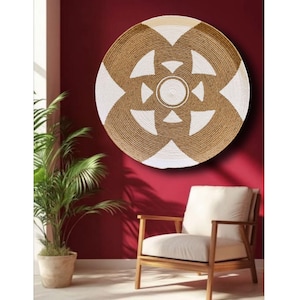 Pode incluir: Uma grande peça de parede redonda com um design tecido em marrom natural e branco. A peça decorativa apresenta uma espiral central e padrões geométricos. Está pendurada numa parede vermelho escuro, com uma cadeira e uma planta em vaso em primeiro plano.