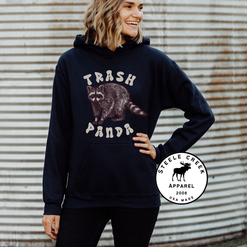 May include: A navy blue hoodie with a raccoon graphic and the text "Trash Panda" in a retro font. The hoodie also features a white circle logo with a moose graphic and the text "Steele Creek Apparel 2008 USA Made".
