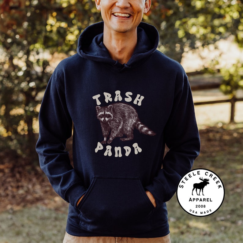 May include: A navy blue hooded sweatshirt with a graphic of a raccoon and the text "Trash Panda". The sweatshirt has a kangaroo pocket and a small logo on the bottom right corner that says "Steele Creek Apparel 2008 USA Made".