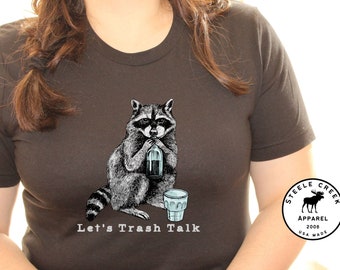 May include: A dark grey t-shirt featuring a black and white illustration of a raccoon holding a bottle and a glass. The text "Let's Trash Talk" is printed below the illustration. The shirt has a round logo on the right sleeve with the text "Steele Creek Apparel 2008 USA Made".