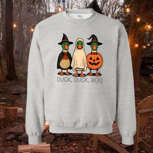 May include: A light gray crewneck sweatshirt featuring a graphic of three ducks dressed for Halloween. One duck wears a witch hat and cape, one is covered by a white ghost sheet, and one is inside a jack-o'-lantern. The text reads DUCK, DUCK, BOO.