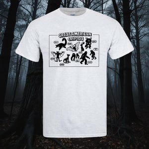 May include: A white t-shirt featuring a black graphic of various cryptids, including the Jersey Devil, Mothman, and Bigfoot. The text "Great American Cryptids" is printed above the graphic.