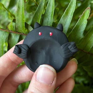 May include: A small, circular, matte black clay sculpture shaped like a bat. The object features two pointed ears at the top, two small red eyes, and two textured wings extending from the sides. The center of the circular body is slightly indented.