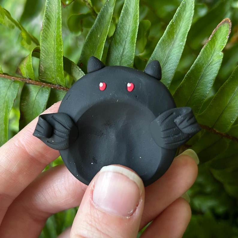 May include: A small, circular, matte black clay sculpture shaped like a bat. The object features two pointed ears at the top, two small red eyes, and two textured wings extending from the sides. The center of the circular body is slightly indented.