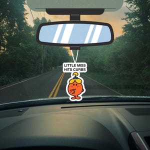 May include: A car interior view with a rearview mirror and a Little Miss car air freshener. The air freshener is orange with a cartoon character design and the text "LITTLE MISS HITS CURBS". The background shows a road and trees.