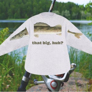 May include: A light gray long-sleeved shirt featuring a detailed illustration of a fish, with the text "that big, huh?" printed below the fish. The shirt is set against a backdrop of a fishing rod and a natural outdoor scene.