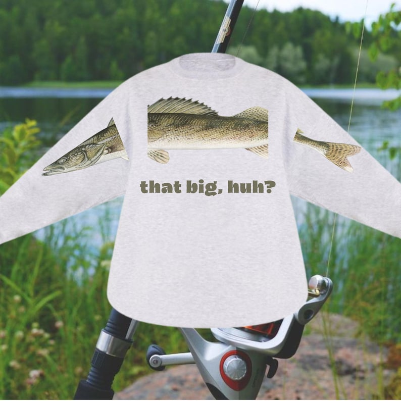 May include: A light gray long-sleeved shirt featuring a detailed illustration of a fish, with the text "that big, huh?" printed below the fish. The shirt is set against a backdrop of a fishing rod and a natural outdoor scene.