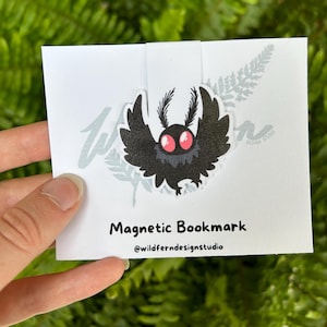 May include: A white card with a black and red moth design magnetic bookmark. The bookmark has the text "Magnetic Bookmark" and "@wildferndesignstudio". The background is a fern.