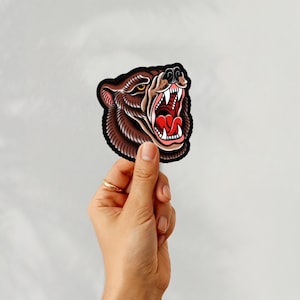 May include: A brown bear head sticker with a black outline, showcasing an open mouth with white teeth and a red tongue. The bear has detailed fur and yellow eyes. The sticker is held by a hand against a white background.