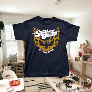 Peut inclure: Un t-shirt bleu marine avec un motif doré et blanc d'un oiseau avec le texte "Smokey and the Bandit East Bound and Down".