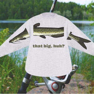 May include: A light gray long-sleeve shirt featuring a fish design on the sleeves and torso. The fish is depicted in shades of green, brown, and black. The text "that big, huh?" is printed below the fish graphic.