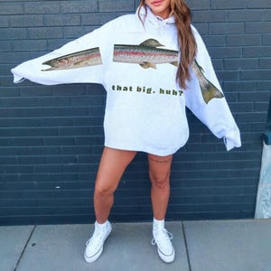 May include: A light gray hoodie with a fish graphic. The hoodie features a large fish illustration across the front and sleeves, with the text "that big, huh?" printed below the fish. The hoodie is paired with white sneakers.