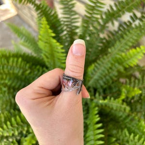 May include: A clear resin ring with a floral design and a silver spoon handle detail. The ring is worn on a thumb, with a backdrop of green fern leaves.