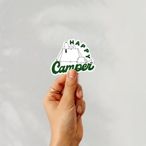 May include: A white sticker with a green outline and text. The sticker features a cartoon illustration of a dog in a tent with the words "Happy Camper" in green. The sticker is held by a hand with a gold ring.