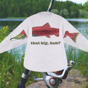 May include: A light gray long-sleeve sweatshirt featuring a red fish illustration with the text "that big, huh?" The fish design is split across the sleeves and torso. The background shows a fishing rod and a lake.