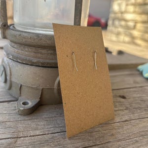 May include: A brown earring display card with two silver earring hooks. The card is made of cardboard and is displayed on a wooden surface. The background includes a vintage metal lamp and a glimpse of an outdoor setting.