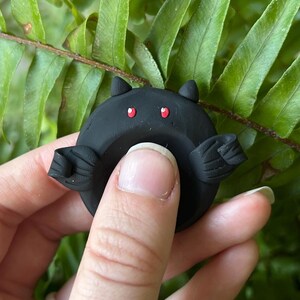 May include: A small, circular black clay figure shaped like a bat with two pointed ears, two small red eyes, and two textured wings extending from the sides. The object is matte black and features a smooth, rounded surface with a central indentation.
