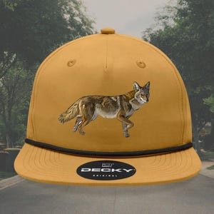 May include: A mustard yellow baseball cap with a black brim and a black rope detail. The cap features a coyote graphic and the text "DECKY ORIGINAL" on the front.