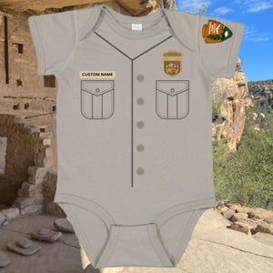 May include: A light gray baby onesie designed to resemble a park ranger uniform. It features a "CUSTOM NAME" tag, two faux pockets, and a National Park Service patch. The onesie has a button-down design and short sleeves.