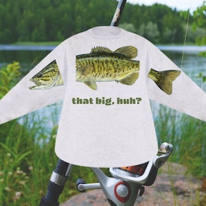 May include: A light gray long-sleeve shirt featuring a large fish graphic with green and yellow details. The text "that big, huh?" is printed below the fish. The shirt is set against a blurred outdoor background.