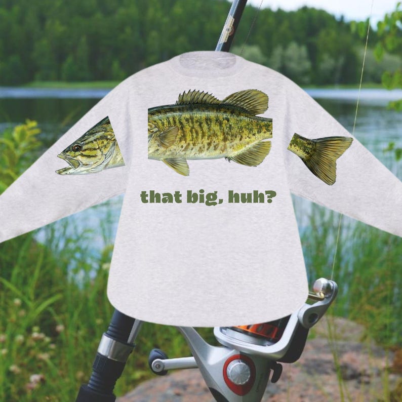 May include: A light gray long-sleeve shirt featuring a large fish graphic with green and yellow details. The text "that big, huh?" is printed below the fish. The shirt is set against a blurred outdoor background.