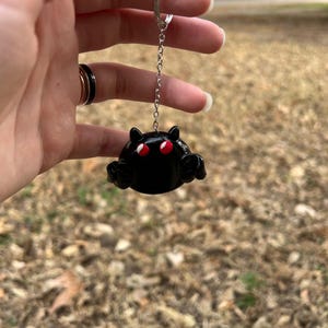 May include: A black keychain featuring a rounded creature with two small pointed ears, two bright red eyes, and two small arms extended forward. The figure is attached to a silver-toned metal chain and a circular split ring for attachment.