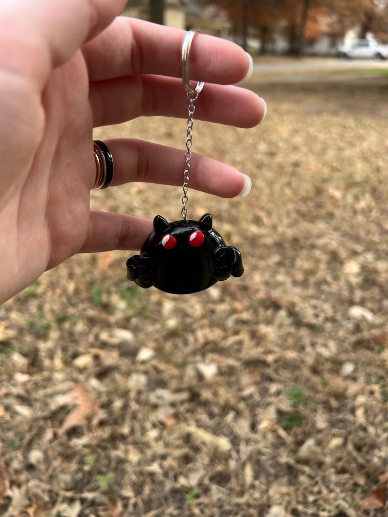 May include: A black keychain featuring a rounded creature with two small pointed ears, two bright red eyes, and two small arms extended forward. The figure is attached to a silver-toned metal chain and a circular split ring for attachment.
