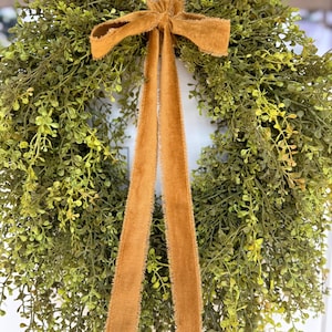 May include: A round, green wreath with small leaves and a brown velvet bow and streamers. The wreath is hanging on a white door. The bow and streamers add a touch of elegance to the natural greenery.