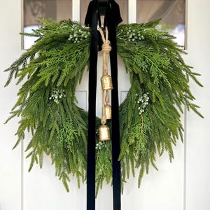 May include: A large, circular evergreen wreath with a lush, textured appearance. The wreath is adorned with small white berries and gold bells hanging from a jute rope. A wide, black velvet ribbon is used to hang the wreath on a white door.