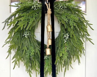 Op de afbeelding: Een grote, cirkelvormige groene krans met een weelderig, gestructureerd uiterlijk. De krans is versierd met kleine witte bessen en gouden bellen die aan een jute touw hangen. Een breed, zwart fluwelen lint wordt gebruikt om de krans op te hangen.