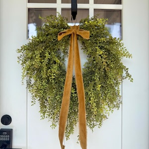 May include: A round, green wreath made of small, leafy branches, adorned with a mustard-colored velvet bow and long, flowing ribbons. The wreath hangs on a white door with a black door handle and a window above.
