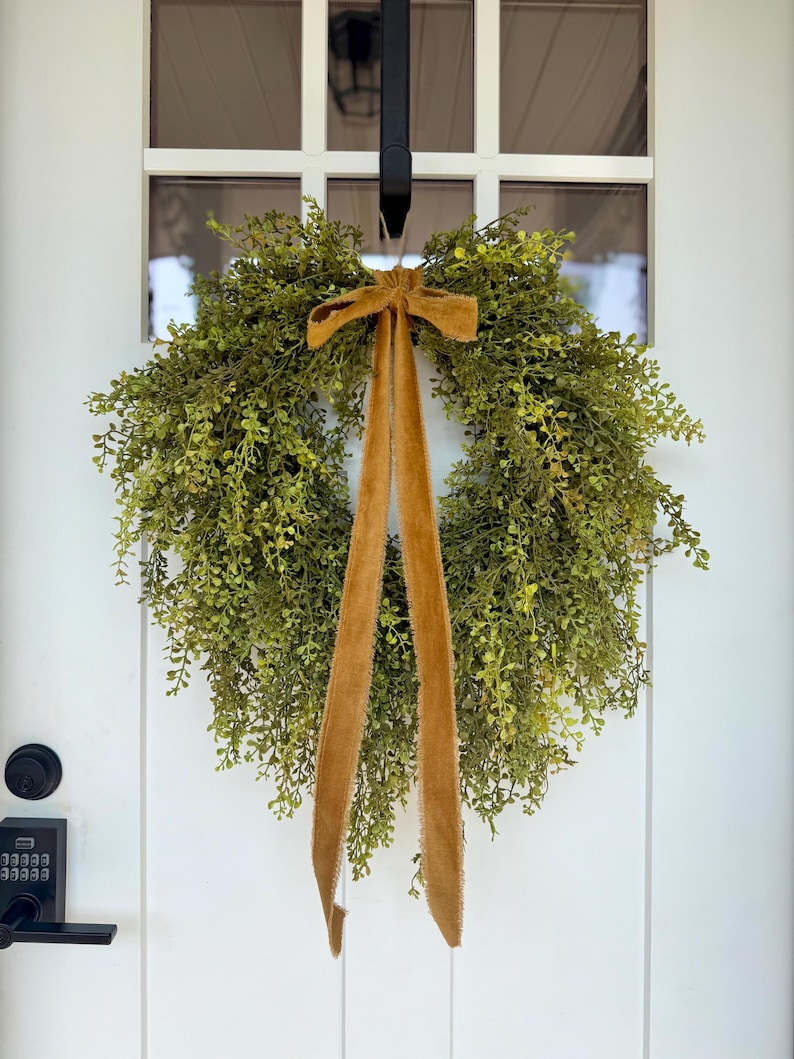 May include: A round, green wreath made of small, leafy branches, adorned with a mustard-colored velvet bow and long, flowing ribbons. The wreath hangs on a white door with a black door handle and a window above.