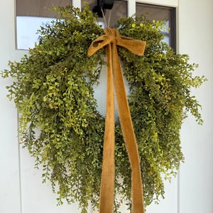 May include: A round, green wreath made of small, leafy sprigs, adorned with a brown velvet bow and long, flowing ribbons. The wreath hangs on a white door, creating a welcoming and natural aesthetic.