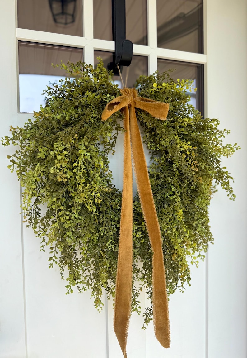 May include: A round, green wreath made of small, leafy sprigs, adorned with a brown velvet bow and long, flowing ribbons. The wreath hangs on a white door, creating a welcoming and natural aesthetic.
