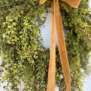 May include: A lush green wreath with delicate leaves and a warm brown ribbon. The wreath is made of faux greenery and is perfect for adding a touch of nature to any home decor.