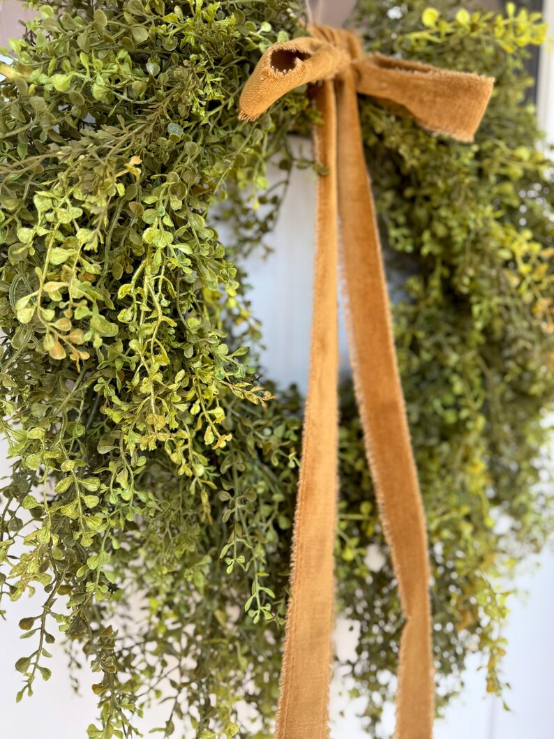 May include: A lush green wreath with delicate leaves and a warm brown ribbon. The wreath is made of faux greenery and is perfect for adding a touch of nature to any home decor.