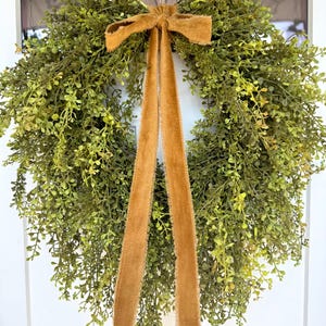 May include: A round, green artificial wreath with small leaves and a brown velvet bow and streamers. The wreath is hanging on a white door, creating a welcoming and natural aesthetic. Perfect for home decor.