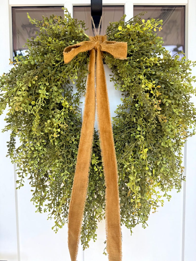 May include: A round, green artificial wreath with small leaves and a brown velvet bow and streamers. The wreath is hanging on a white door, creating a welcoming and natural aesthetic. Perfect for home decor.