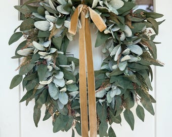 May include: A decorative wreath featuring a mix of green and gray-green leaves, accented with brown sprigs and a gold velvet ribbon. The wreath is designed for door or wall decor.