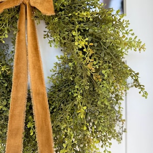 May include: A close-up of a green wreath with small leaves and a mustard-colored velvet ribbon. The wreath is hanging against a white background. The ribbon is tied in a bow at the top and hangs down in two long strands.