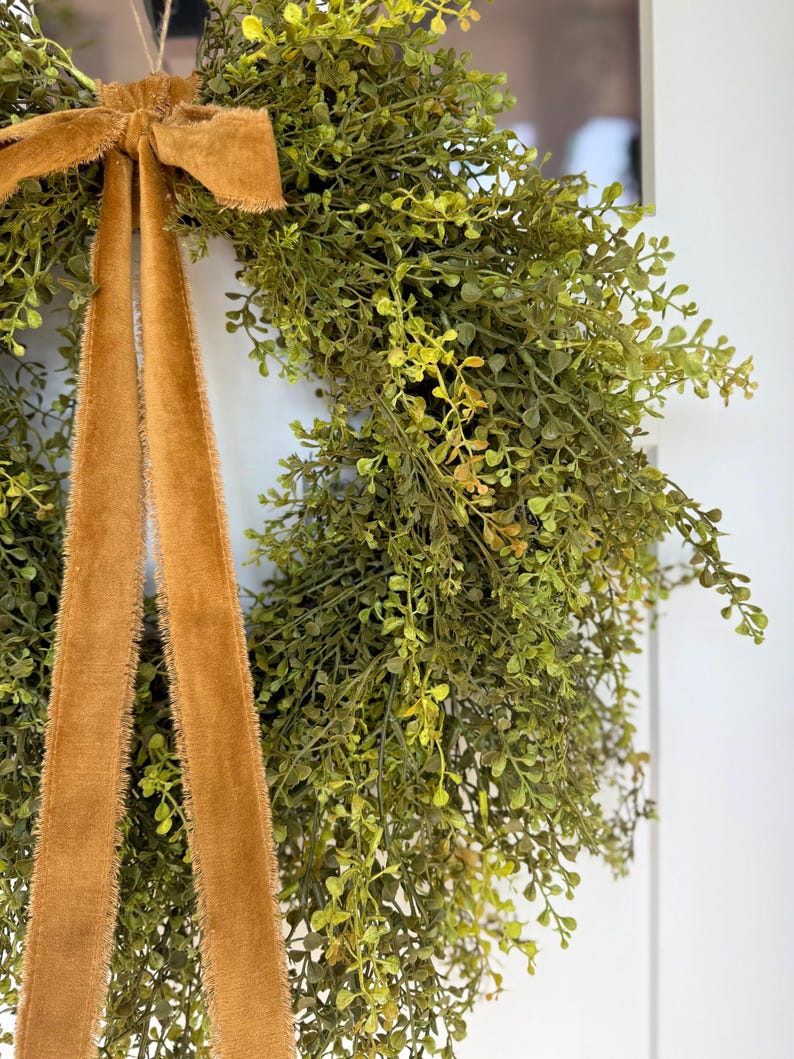 May include: A close-up of a green wreath with small leaves and a mustard-colored velvet ribbon. The wreath is hanging against a white background. The ribbon is tied in a bow at the top and hangs down in two long strands.