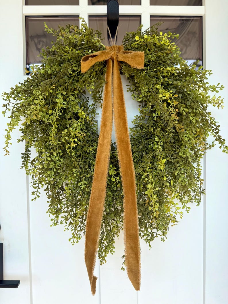 May include: A round, green wreath made of small, leafy sprigs. A long, mustard-colored velvet ribbon is tied in a bow at the top and hangs down. The wreath is hanging on a white door.
