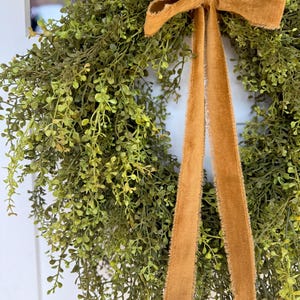 May include: A lush green wreath with small, delicate leaves, accented by a warm, golden-brown velvet bow and long, flowing ribbons. The wreath is designed for home decor and adds a touch of natural elegance.