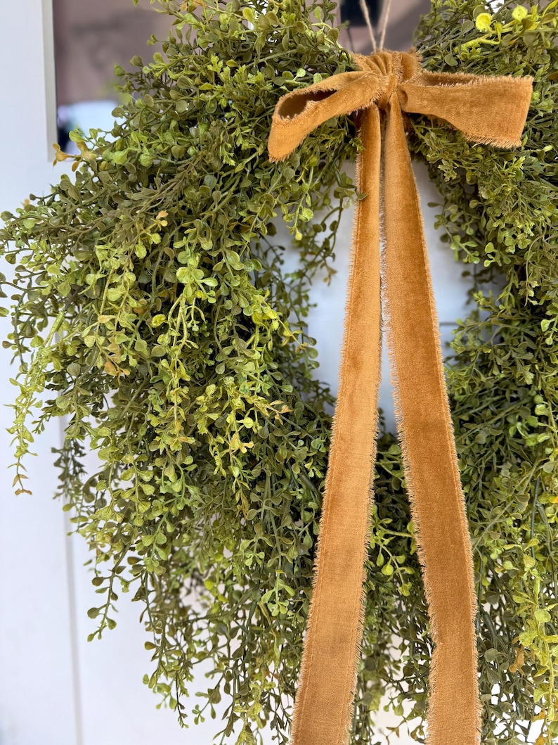 May include: A lush green wreath with small, delicate leaves, accented by a warm, golden-brown velvet bow and long, flowing ribbons. The wreath is designed for home decor and adds a touch of natural elegance.