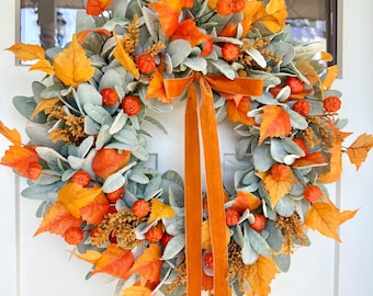 May include: A circular autumn wreath featuring sage green lamb's ear leaves, vibrant orange maple leaves, small orange pumpkins, and golden-brown filler accents. A long, solid orange velvet ribbon hangs from the top center of the wreath.