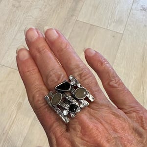 May include: A silver statement ring with an abstract design. The ring features black and grey stones in various shapes, and small clear crystals. The ring is worn on a hand.