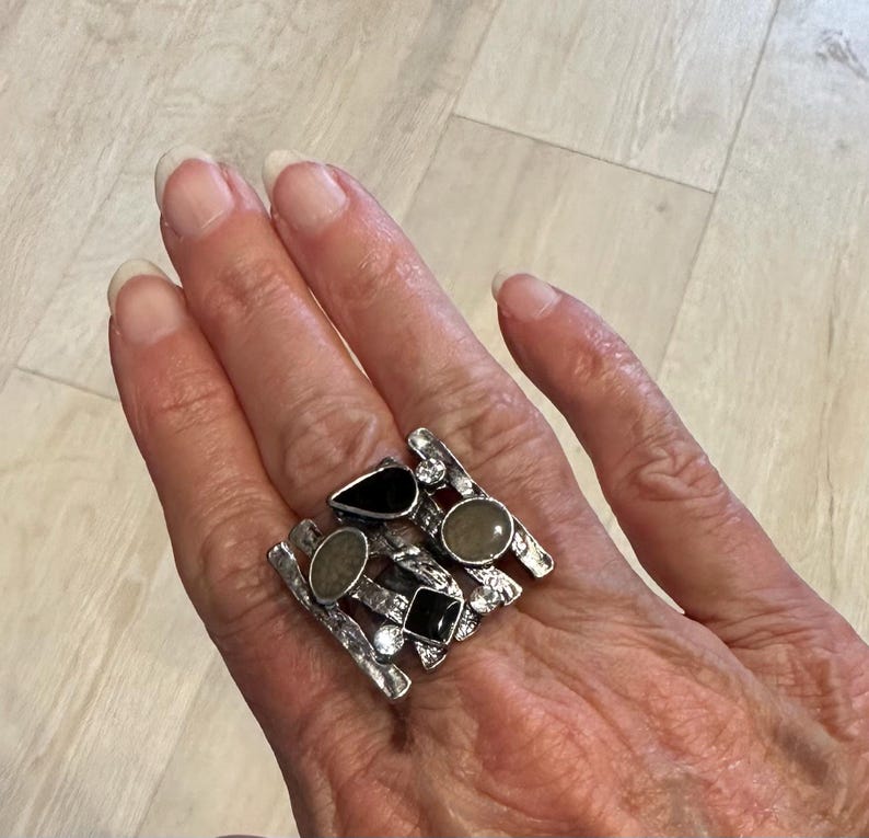 May include: A silver statement ring with an abstract design. The ring features black and grey stones in various shapes, and small clear crystals. The ring is worn on a hand.