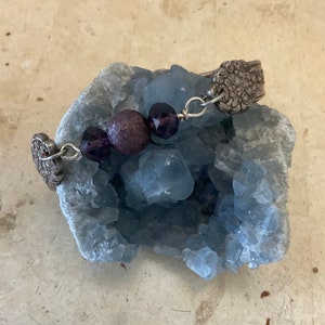 May include: A silver bracelet with a purple bead and two purple glass beads. The bracelet is on a blue crystal rock.